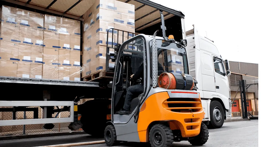 Eficiência e logística 27 Uma empilhadeira de combustão interna, de cor laranja e cinza, está elevando um grande palete de caixas envoltas em plástico para carregamento na parte traseira aberta de um caminhão branco.